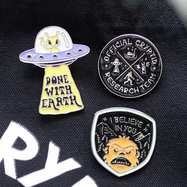 Circular enamel pin depicting four mythological creatures including big foot and mothman. Text reads Official Cryptid Research Team.