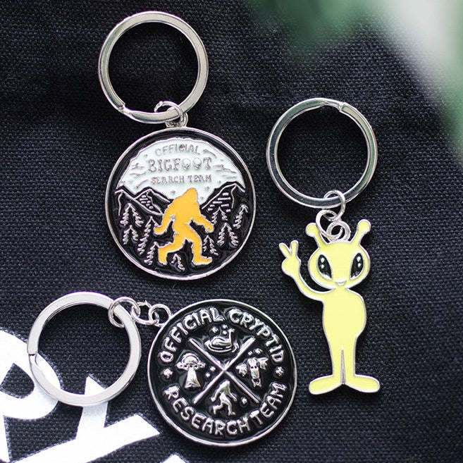 Circular enamel keyring depicting Bigfoot walking through the woods. Text reads Official Bigfoot Search Team. Presented alongside other items from the same range.