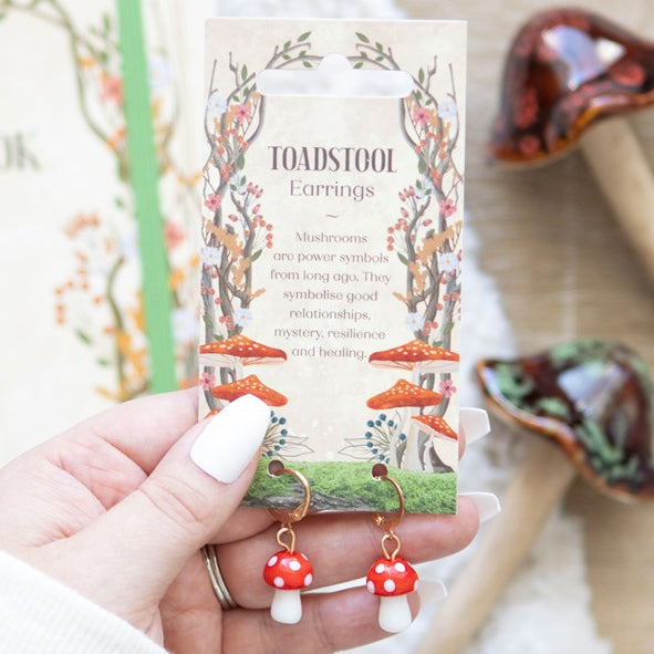 Toadstool shaped earrings, hanging from small gold hoops. Presented on a themed backing card. 