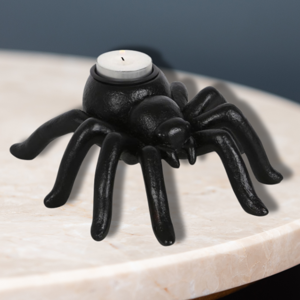 Black resin spider-shaped tea light holder, placed on a circular table with a dark blue background. 