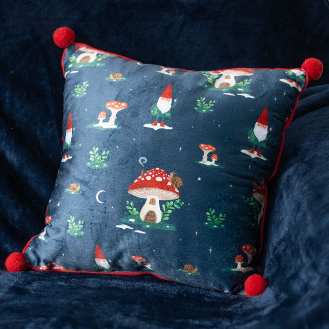 Square decorative pillow with gnome and mushroom-house motif. Dark blue with red pom-pom embellishments on the corners.